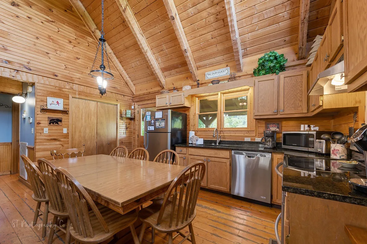 The image features an open kitchen and dining area with exposed wooden beams, a roomy wooden dining table, modern appliances, and large windows. The kitchen is clearly designed for group meal prep and easy social interaction, while the table invites shared experiences.