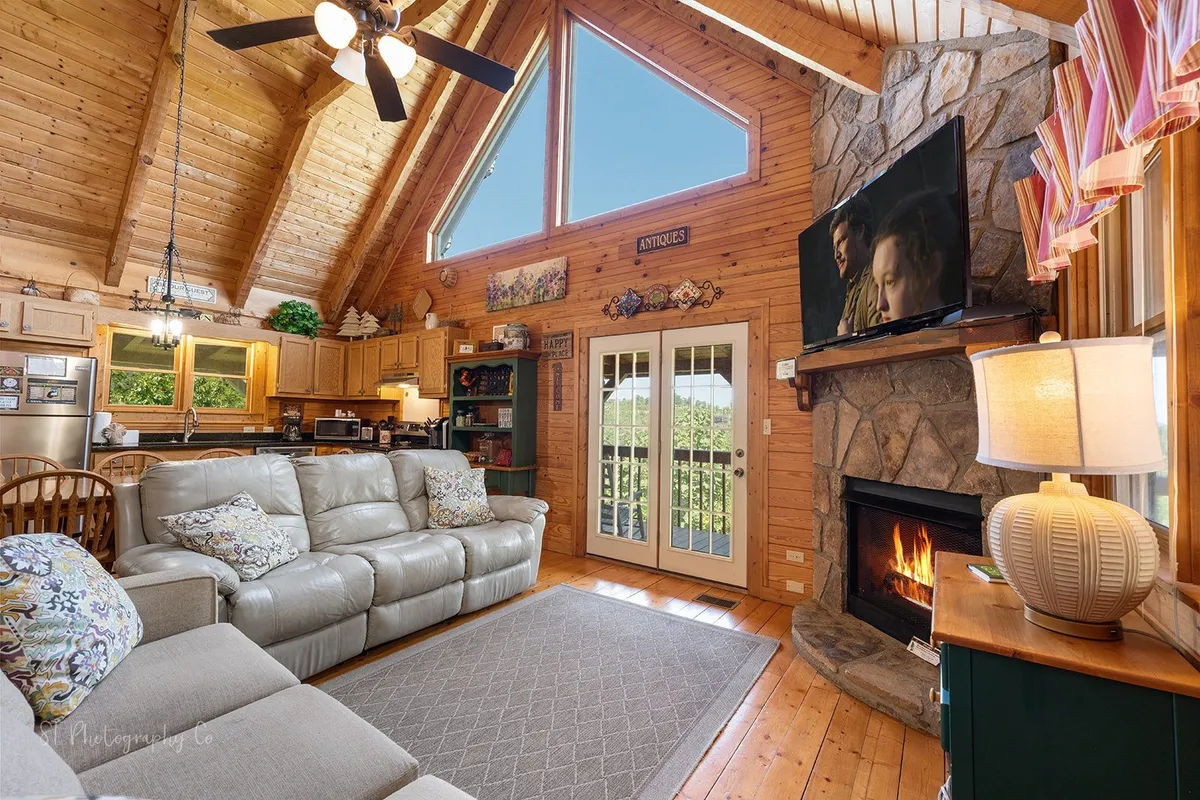 This image showcases a spacious living room with a comfortable leather sectional sofa arranged by a stone fireplace, above which a flat-screen TV is mounted. Large windows offer scenic views and bathe the space in natural light, creating a cozy yet open atmosphere ideal for family gatherings or movie nights.