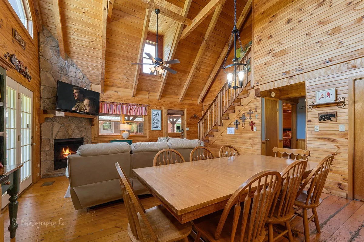 A spacious, open-plan living and dining area featuring a large wooden dining table, comfortable sofa, stone fireplace, and staircase. The space exudes warmth, perfect for gathering after a day exploring the Smoky Mountains and local attractions.