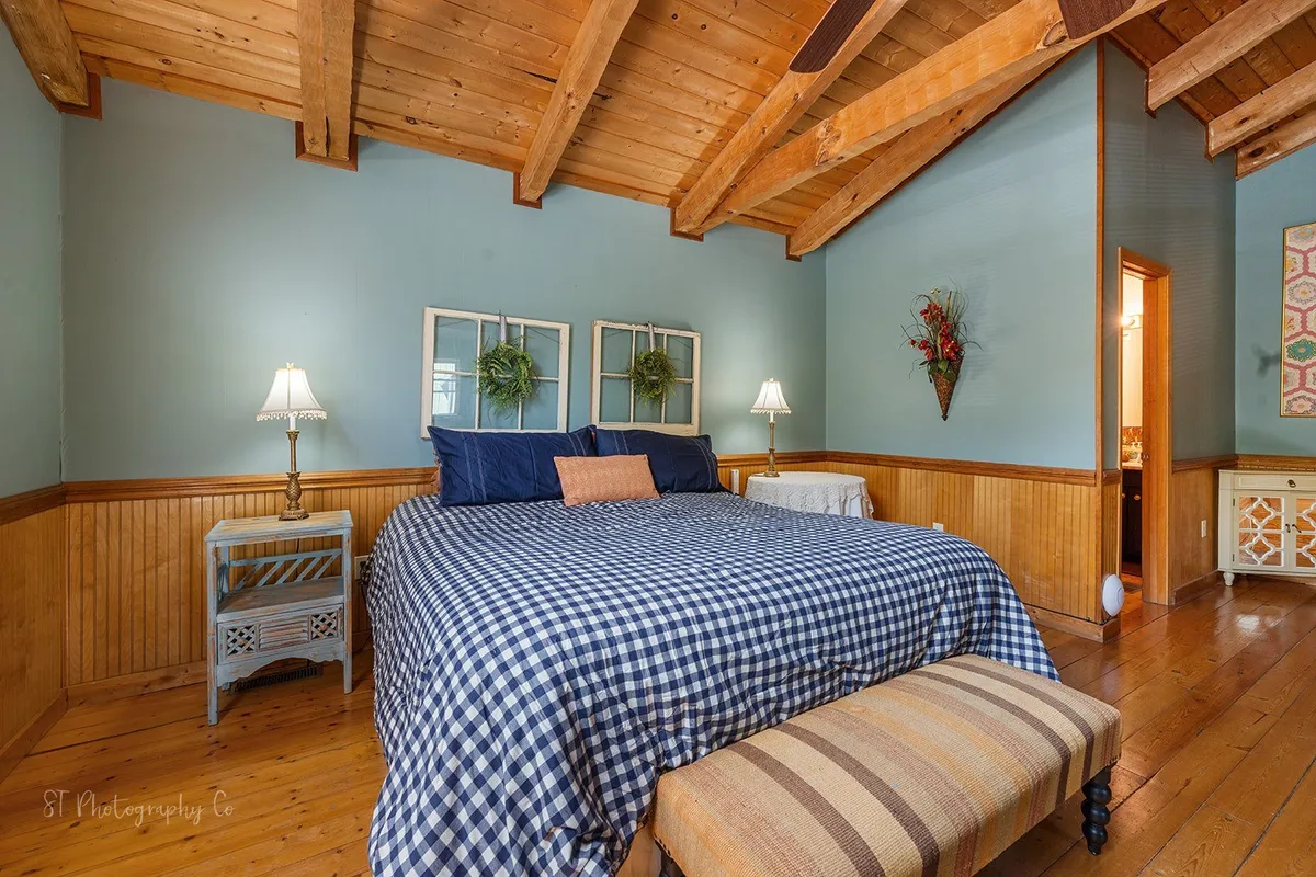 A warmly lit bedroom features a bed with inviting plaid bedding, rustic wooden beams overhead, a nightstand with a lamp, and decorative wall art. Natural light flows into the space, creating a serene and comfortable atmosphere perfect for relaxation and unwinding after a busy day.