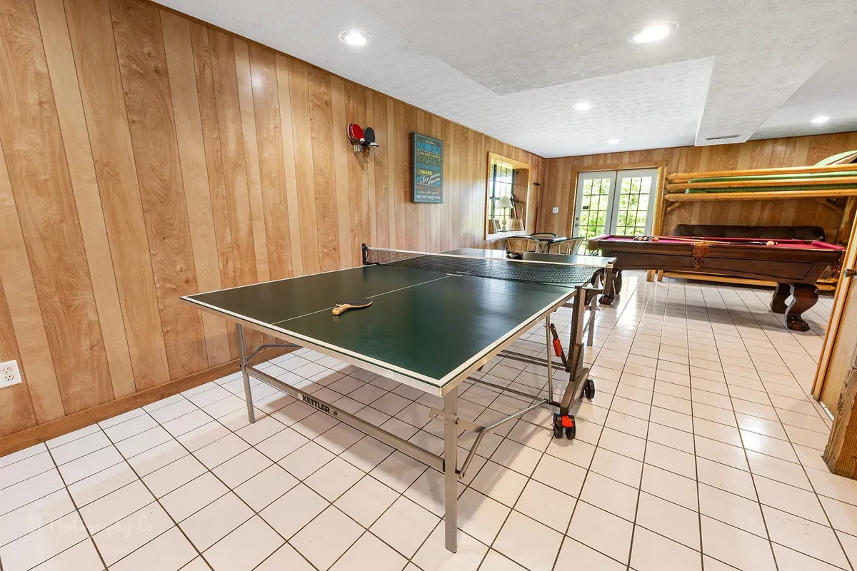 This image captures a lively, wood-paneled game room featuring both a ping pong table and a pool table. Natural light filters in through the windows to illuminate the recreational space, which is designed for friendly competitions and shared fun.