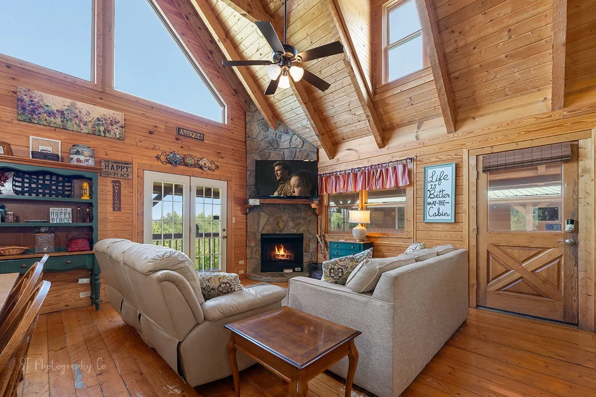A spacious, open-concept living area with a stone fireplace, ample comfortable sofa seating, and floor-to-ceiling windows that fill the space with natural light. Wooden beams and decorative wall art add rustic charm, while the arrangement encourages gathering and relaxation. Nature is visible through the windows, subtly connecting the indoors to the scenic outdoors.