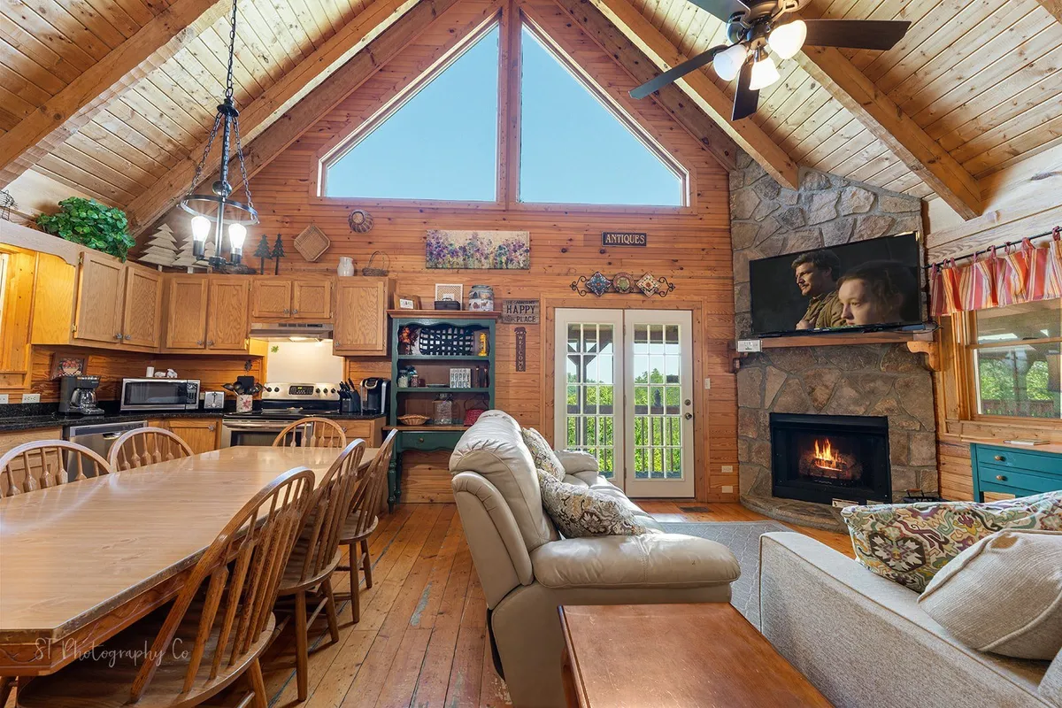 A large wooden dining table sits adjacent to a stone fireplace, surrounded by comfortable seating and illuminated by natural light streaming through generously sized windows. The space is designed for communal dining, socializing, and enjoying mountain sunsets together.