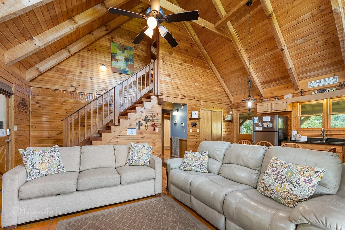 A cozy and inviting open-plan living space with comfortable sofas, rustic wooden beams, a visible staircase, and a glimpse of the kitchen area. Thoughtful decorative touches and artwork create a welcoming environment perfect for family gatherings and relaxation after a day out.