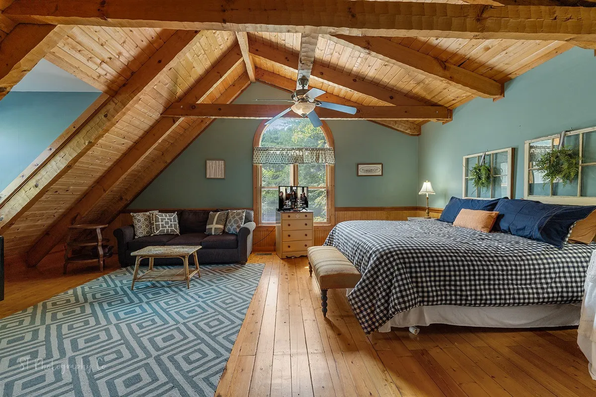 A spacious bedroom with a large bed, cozy sofa, decorative rug, and window that opens to scenic views. The wooden ceiling beams and soft lighting add to the inviting, tranquil feel of the space, emphasizing comfort and flexibility for both relaxation and social moments.