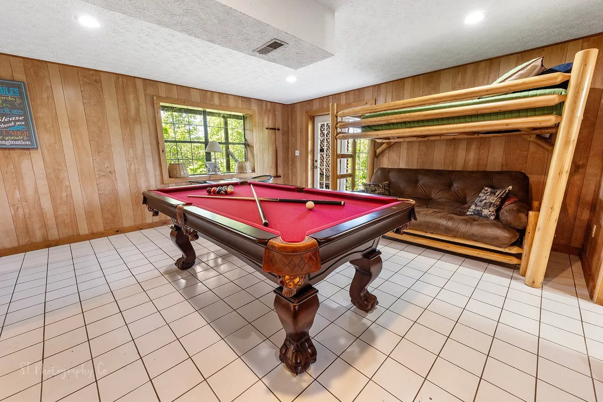 A playful, relaxed game room featuring a prominent pool table, a twin-over-futon bunk bed, a sofa, and generous windows letting in daylight. The arrangement shows how the space doubles as both a recreation and sleeping area.