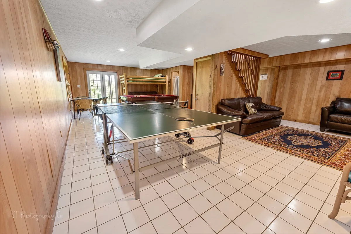 This image displays a vibrant game room featuring a ping pong table at the center, a comfortable leather sofa against one wall, and a staircase leading to another level. The space is set up for both active play and relaxed lounging, offering a lively, inviting atmosphere ideal for families and friends.