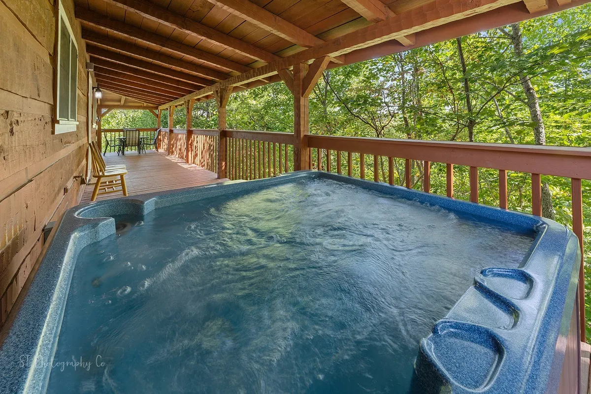 A spacious wooden deck nestled among mature trees, featuring a bubbling hot tub as the focal point. The hot tub sits invitingly with comfortable seating nearby, perfect for relaxing while immersed in nature. The deck is surrounded by lush forest, offering privacy and a tranquil ambiance, with plenty of room for gathering and socializing.