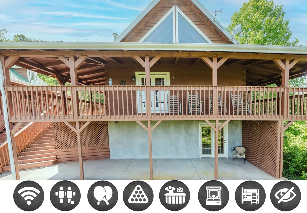 The image shows the exterior of a charming cabin with a spacious wooden balcony, large triangular windows, and a green lawn area. The balcony is positioned to capture sweeping mountain sunset views, offering a peaceful space for guests to relax, enjoy coffee, or gather with loved ones while surrounded by nature.