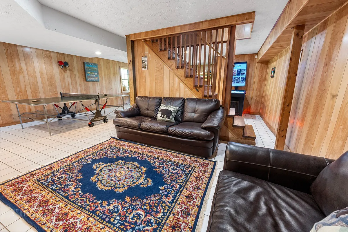 A spacious and comfortable game room featuring a large sofa, plush rug, and a table tennis table. The room is filled with natural light and has an inviting atmosphere for relaxing, playing, and socializing. Stairs lead up to another area of the cabin, emphasizing the open and family-friendly layout.