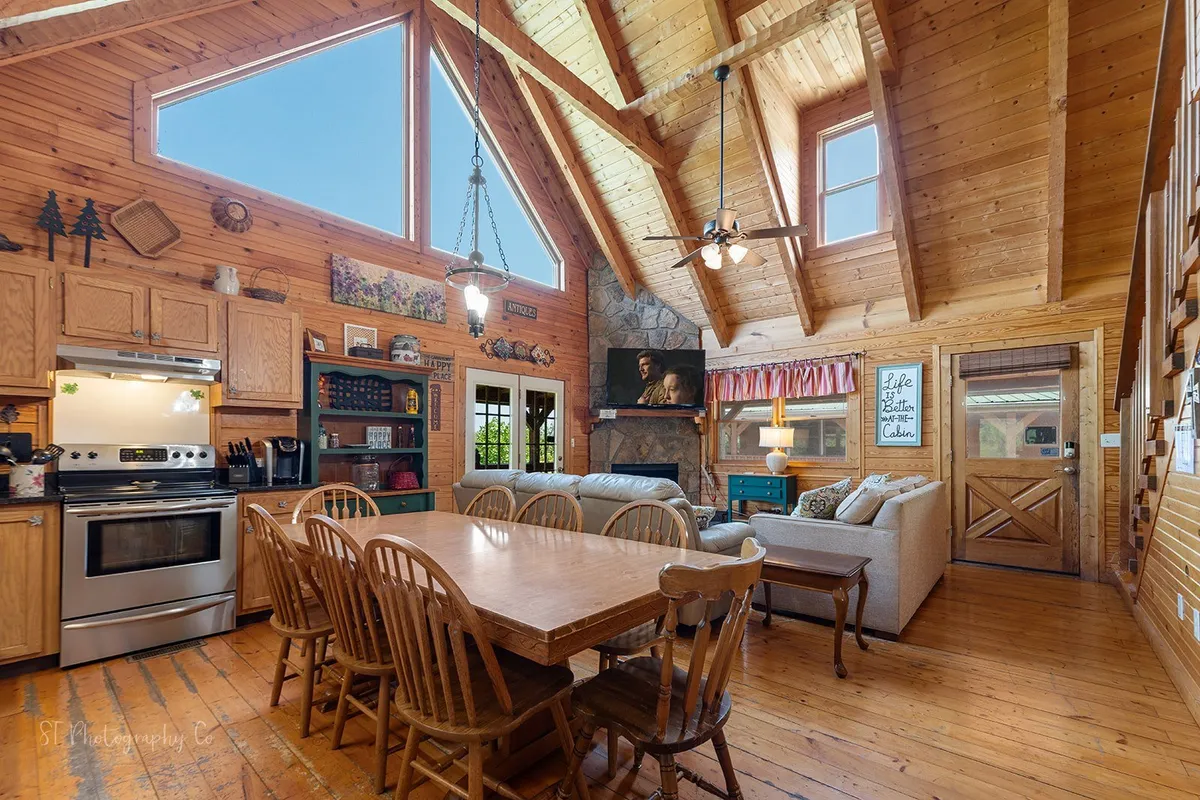 This interior scene captures the open-plan layout with a large wooden dining table, a stone fireplace, comfortable seating, expansive windows, and visible kitchen appliances. The setting exudes warmth and togetherness, perfect for family meals, celebrations, or relaxed entertaining.